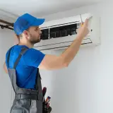 Technik w niebieskiej czapce i szarym ogrodniczkach naprawia klimatyzator śrubokrętem.
