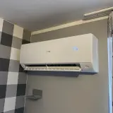 Biała jednostka klimatyzacyjna Haier zamontowana na szarej ścianie, obok wzorzystej tapety i zasłon.