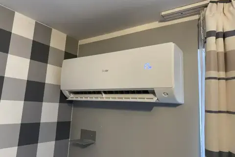 Biała jednostka klimatyzacyjna Haier zamontowana na szarej ścianie, obok wzorzystej tapety i zasłon.